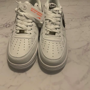 Nike Air Force 1 vit/svart sneakers - Säljer ett par klassiska Nike Air Force 1 sneakers i vitt skinn med svart swoosh. Skorna har perforerad tå, platt sula och snörning. Ikonisk design med metallmärke på snörena och tydlig Air-detalj på sulan. Perfekta för dig som gillar streetstyle och tidlös look.