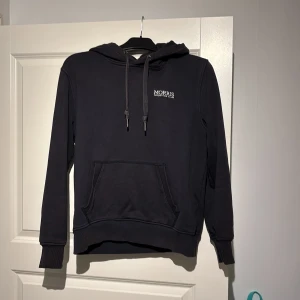 Svart hoodie från Morris - Svart hoodie från Morris med vit broderad logga på bröstet. Klassisk känguruficka framtill, justerbar huva med snörning och ribbade muddar vid ärmslut och nederkant. Perfekt för en chill och stilren look. Ej fast pris så kom med bud! (den är marinblå/svart)