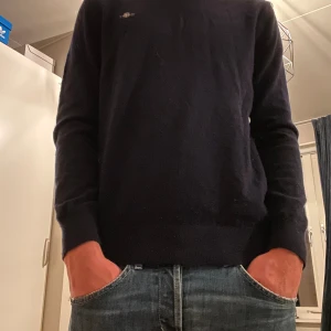 Mörkblå stickad tröja med diskret logga - Mörkblå stickad tröja med lång ärm och rund hals. Tröjan har en liten, diskret logga på bröstet och är enkel i modellen, perfekt för en clean och stilren look. Passar dig som gillar klassiska basplagg med en snygg finish.