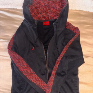 Svart/röd kofta från Hugo - Svart hoodie från Hugo med dragkedja och huva. Rött abstrakt mönster på huva och längs ärmarna ger en unik look. Perfekt för dig som vill sticka ut. Skön passform och snygga detaljer som röda snören vid huvan.