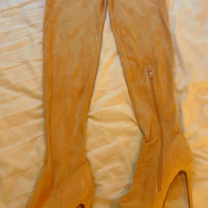 Beiga höga stövletter med klack - Snygga höga stövletter i beige mocka med dragkedja på sidan och hög klack. Skorna har en plattform framtill och en spetsig tå, vilket ger en trendig och elegant look. Perfekta för att lyfta vilken outfit som helst.