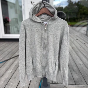 Grå hoodie med dragkedja och huva - Mysig grå hoodie med dragkedja framtill och två fickor. Tröjan har huva med snörning och är långärmad. Perfekt för chill dagar eller när du vill ha något bekvämt och stilrent. Materialet är kashmir så super skönt och mjukt.