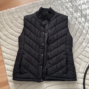 Svart quiltad dunväst från Joy - Svart quiltad dunväst från Joy med hög ribbad krage och både dragkedja och tryckknappar framtill. Västen har två fickor med dragkedja och är tillverkad i 100% polyester. Perfekt att slänga över en hoodie eller tröja när det är kyligt ute.