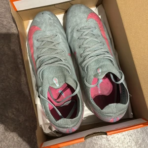 Nike fotbollsskor grå med rosa detaljer modell är PRO Nike  - Säljer ett par grå Nike fotbollsskor med snygga rosa swoosh-detaljer och turkosa dobbar. Skorna har snörning och är designade för att ge bra grepp på planen. Perfekta för dig som vill sticka ut med stil på matchen.