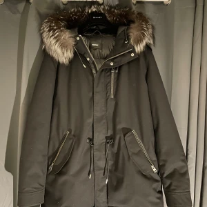 Svart Mackage parkasjacka med päls - Snygg svart parkasjacka från Mackage med fluffig päls runt huvan (går att ta av) och quiltad dunfyllning. Jackan har dragkedjor på fickorna, snörning i midjan och en varm, glansig insida. Perfekt för kalla vinterdagar och riktigt stilren look