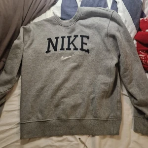 Grå sweatshirt från Nike - Klassisk grå sweatshirt från Nike med stor svart logga och broderad swoosh på bröstet. Tröjan har rund halsringning, ribbade muddar och är tillverkad i mjuk bomullsblandning. Perfekt för en avslappnad och sportig stil.