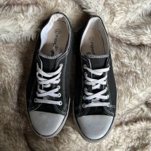 Svarta låga sneakers med vit sula - Klassiska svarta sneakers i canvas med vita snören och vit gummisula. Skorna har rund tå, kontrastsömmar och lågt skaft. Lite fläckar på sulan.