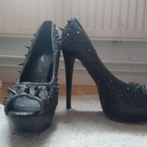 Svarta pumps med nitar från Belle Women - Riktigt snygga svarta pumps med hög klack och platåsula från Belle Women. Skorna har öppen tå, är täckta av svarta nitar och glittrande detaljer som ger en edgy vibe. Perfekta för dig som vill sticka ut och älskar statementskor.