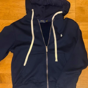 Marinblå hoodie från Polo Ralph Lauren - Snygg marinblå hoodie från Polo Ralph Lauren med dragkedja, vita snören och klassisk logga på bröstet. Tröjan har huva, fickor framtill och är tillverkad i mjuk bomullsmix. Perfekt för chill dagar eller när du vill ha en clean streetstyle.