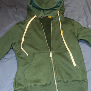 Grön hoodie från Polo Ralph Lauren - Mörkgrön hoodie från Polo Ralph Lauren med dragkedja, huva och vita snören. Broderad gul logga på bröstet och mjuk fleece på insidan. Perfekt för kyliga dagar och har klassiska kängurufickor framtill.