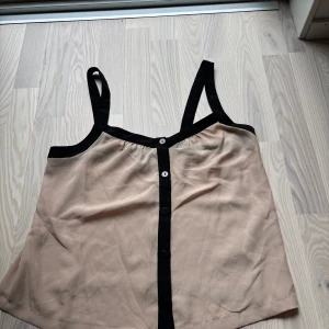 Beige linnetopp - Säljer en beige linnetopp från HM Divided med svarta kontrastdetaljer och knappar framtill. Toppen har breda svarta axelband och ärmlös design. Materialet känns lätt och svalt mot huden.