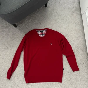 Röd v-ringad tröja från GANT - Röd v-ringad tröja från GANT med broderad logga på bröstet. Tröjan har långa ärmar och klassisk passform. Insidan av kragen har ett rutigt tyg i rött, vitt och blått som ger en snygg detalj. Perfekt för dig som gillar preppy stil.