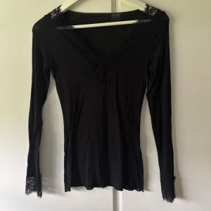 Svart spetsblus från Intimissimi - Svart långärmad blus från Intimissimi med v-ringning och fina spetsdetaljer vid halsringning, axlar och ärmslut. Tunn och mjuk kvalitet med inslag av kashmir för extra lyxig känsla. Perfekt för dig som gillar stilrena och feminina plagg.