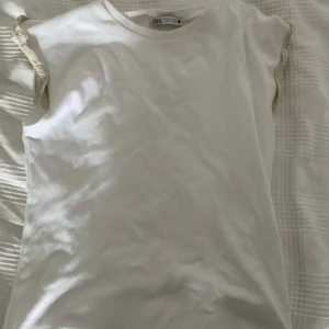 Vit topp med volang från Zara - Säljer en vit topp från Zara med volangdetaljer i beige längs ärmöppningarna. Toppen har rund halsringning och är tillverkad i mjuk bomull, perfekt för en fräsch och enkel look. Är i storlek M men skulle säga att det passar S också💗