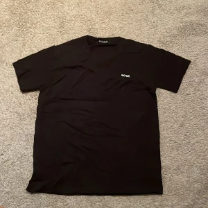 Svart Hugo Boss t-shirt  - Svart t-shirt från Hugo Boss med vit BOSS-logga på bröstet. Klassisk rund hals och korta ärmar. T-shirten har en clean och stilren look, perfekt för dig som gillar enkla designer med premiumkänsla. Materialet är mjuk  som känns skönt mot huden. Den är nästan aldrig använt jag har bara testat den 
