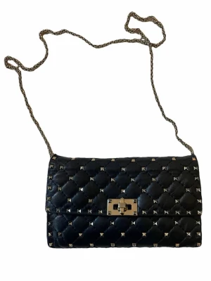 Svart quiltad axelväska Valentino - Säljer en svart quiltad axelväska från Valentino med silverfärgade nitar och kedjerem. Väskan är i skinn och har ett rött innerfack med dragkedja. Stängs med ett snyggt metallspänne framtill och har en lyxig vibe.