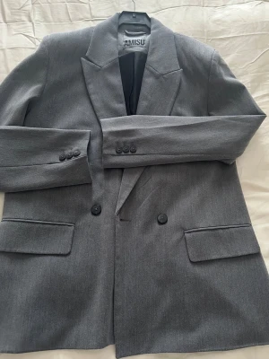 Grå kavaj från Amisu - Säljer en stilren grå kavaj från Amisu med klassisk passform. Kavajen har två knappar framtill, breda slag och fickor med lock. Perfekt för dig som vill ha en enkel men snygg look. Fodrad insida och diskreta knappar vid ärmslut.