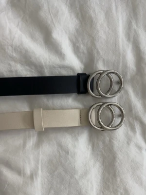 Två stilrena bälten  - Säljer två bälten, ett svart och ett beige, båda med snygga silverfärgade dubbelringar som spänne. Bältena är smala och har en clean look som passar till jeans eller kjol. båda för 40kr eller en för 20kr!!🤍