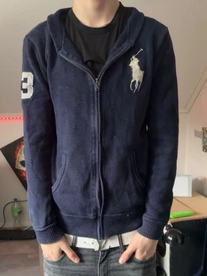 Mörkblå stickad hoodie från Polo Ralph Lauren - Säljer en mörkblå stickad hoodie från Polo Ralph Lauren i storlek S . Tröjan har dragkedja, huva och två fickor framtill. Stor vit logga på bröstet och en vit siffra på ärmen ger en sportig vibe. Perfekt för dig som gillar klassisk streetwear.