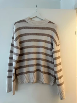 Randig beige/brun stickad tröja Kappahl - Mysig stickad tröja från Kappahl i beige och brunt med breda horisontella ränder. Tröjan har ribbade muddar vid ärmslut och nederkant samt en rundad, ribbad halsringning. Perfekt för lager på lager under kyliga dagar.