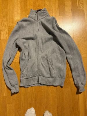 Grå stickad kofta från Polo Ralph Lauren - Säljer en grå stickad kofta från Polo Ralph Lauren med dragkedja framtill och två fickor. Klassisk ribbad krage och muddar, samt den ikoniska mörkblå loggan broderad på bröstet. Perfekt till lager på lager under höst och vinter.