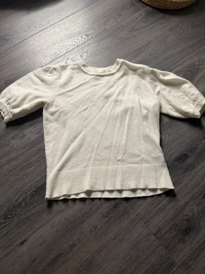 Vit stickad tröja med puffärm - Säljer en vit stickad tröja med korta puffärmar och bred ribbad kant nedtill. Tröjan har en rund halsringning och är gjord i ett mjukt material som passar perfekt till både jeans och kjol. Enkel och stilren design som funkar året runt.