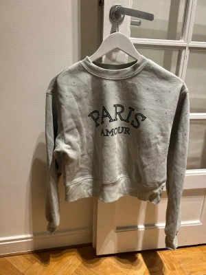 Grå/grön  croppad sweatshirt Paris Amour - Grå croppad sweatshirt med texten 'PARIS AMOUR' framtill. Tröjan har långa ärmar och är dekorerad med små silverfärgade pärlor över hela tyget. Mjuk och skön bomullskänsla, perfekt för en chill look.