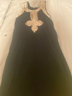 Svart ärmlös klänning med guldmönster - Snygg svart klänning med ärmlös design och dekorativa guldiga detaljer runt halsringningen och mitt fram. Klänningen har en rak passform och ett unikt mönster som ger en cool touch. Perfekt för dig som vill sticka ut lite extra.