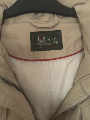Beige vindjacka från Peak Performance - Säljer en beige vindjacka från Peak Performance i storlek L. Jackan har fyra fickor framtill med lock och knappar, dragkedja och hög krage. Perfekt för blåsiga dagar och enkel att matcha med olika outfits.