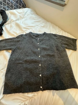 Mörkgrå kofta med pärlknappar - Mysig mörkgrå kofta i stickad ullmix med raka trekvartsärmar och rund hals. Stängs med vita pärlknappar framtill. Perfekt för lager på lager under kyliga dagar.