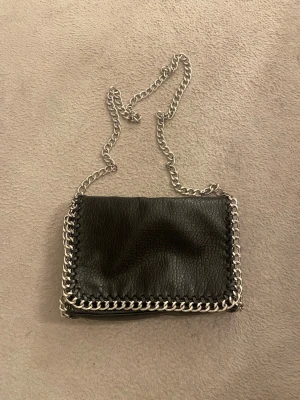 Tiamo väska - Säljer en svart axelväska från Tiamo i skinnimitation med grov silverfärgad kedja som axelrem och detaljer runt kanterna. Väskan har magnetlås och ett innerfack med dragkedja. Perfekt storlek för det viktigaste och ger en cool vibe till din outfit.