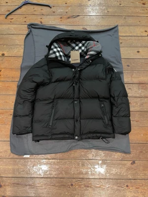 Svart pufferjacka från Burberry - Säljer en svart pufferjacka från Burberry med klassiskt rutigt foder i huvan. Avtagbara armar för svalare väder. Storlek: Large, men kan justeras till Medium. Jackan är varsamt använd och taggen är påsatt för bildernas skull. Priset kan diskuteras och den skickas så snabbt om möjligt.