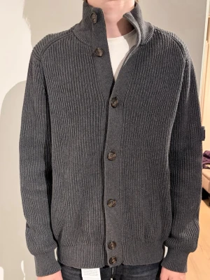 Grå stickad kofta från Henri Lloyd - Säljer en grå ribbstickad kofta från Henri Lloyd med knappar framtill och ett snyggt patch med brittiska flaggan på ärmen. Koftan har lång ärm, normal passform och är gjord i mjukt material som passar perfekt till kyliga dagar.