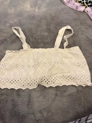 Vit broderad väst med knappar - Säljer en söt vit väst i bomull med broderade blommönster och spetsdetaljer. Västen har smala axelband och knäpps med små knappar framtill. Perfekt till sommaren för en romantisk och luftig look.