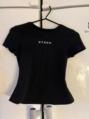 Svart topp från GTECH - Säljer en svart topp från GTECH med korta ärmar och rund hals. Toppen har en slim passform och är tillverkad i mjukt bomullsmaterial. Enkla vita bokstäver på bröstet ger en clean och sportig look.