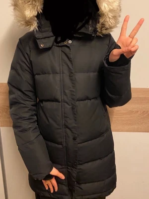 Mount Everest puffjacka med päls - Säljer en svart lång pufferjacka med huva och beige fuskpäls. Jackan har dragkedja och knappar framtill, samt quiltad design för extra värme. Perfekt för kalla dagar och riktigt skön att bära. Passar dig som vill ha en stilren och varm vinterjacka.