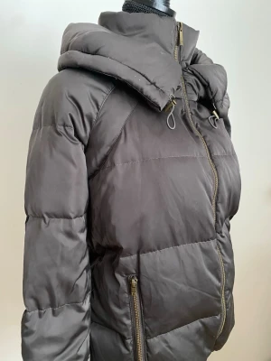 Glansig pufferjacka med hög hals  - Riktigt snygg pufferjacka i mörkgrön/brun färg. Två fickor med dragkedja på båda sidorna, resår längst ner och slutet på ärmarna. Jackan har inte luva, utan extra lager för att hålla nacken/halsen varm! 🌺  ‼️OBS. Finns en skada högre upp på tyget bakom dragkedjan. Går att stänga hela vägen, men försiktigt så den inte fastnar! 🙌 (skada syns på tredje bild) ‼️  🪡 Storlek: XS  📏 Material: 100% polyester  ✉️ Kontakta vid frågor! 