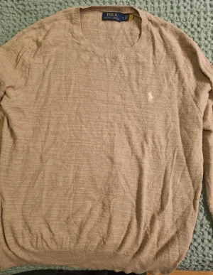 Beige stickad tröja Polo Ralph Lauren - Beige stickad tröja från Polo Ralph Lauren i storlek M. Tröjan har rund halsringning, diskret struktur i materialet och den klassiska broderade loggan på bröstet.