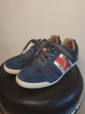 Pantofola d’Oro blå sneakers - Säljer ett par riktigt snygga sneakers från det anrika italienska märket Pantofola d'Oro. Skorna har en sportig men lyxig look i en djupt mörkblå färg med de klassiska italienska färgdetaljerna på sidan och undersidan.  Riktiga skinnskor från Italien!   Färg: Marinblå (Navy) med detaljer i rött, vitt och grönt.  Design: Snygga metalldetaljer i form av stjärnor på sidorna och broderad logotyp på plösen och hälen.  Skick: Mycket bra begagnat skick! De är väl omhändertagna och har mycket kvar att g