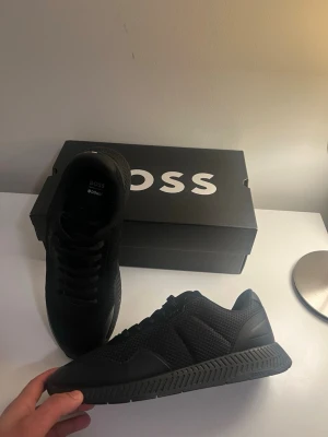 Svarta sneakers från BOSS - Snygga helsvarta sneakers från BOSS med meshdetaljer och stilren design. Skorna har snörning, rund tå och en rejäl sula med diskret BOSS-logga på sidan. Perfekta för dig som gillar en clean och modern look.