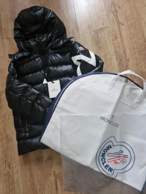 Moncler pufferjacka - Säljer en svart pufferjacka från Moncler med blank finish och stor huva. Jackan har dragkedja framtill, Moncler-logga på ärmen och coola vita detaljer på ena armen. Perfekt för kalla dagar och riktigt snygg streetstil. Köpte i Manchester oktober 22 2025. Orginal pris £980. Väska och påse yill förpackning är jnkluderade. Storlek 4 eller XL till L. Skana QR kodeb eller logon på vänstra armen för att checka autentisering