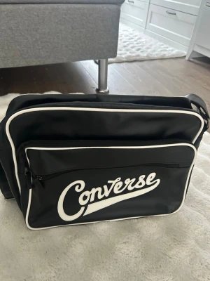 Svart axelväska från Converse - Säljer en svart axelväska från Converse med vit logga och vita detaljer. Väskan har justerbar axelrem, stort huvudfack och en framficka med dragkedja. Klassisk retrostil och tillverkad i syntetmaterial.
