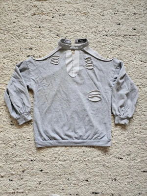 Grå sweatshirt med cut out-detaljer - Grå sweatshirt med hög hals och knapp i nacken, cut out-detaljer framtill och på axlarna samt långa ärmar med breda muddar. Tröjan har en loose passform och är gjord i mjuk bomullsmix. Perfekt för dig som gillar edgy streetstyle.