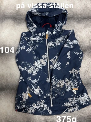 Blommig marinblå vindjacka från Reima - Snygg marinblå vindjacka från Reima med vitt blommönster. Jackan har huva, dragkedja framtill och praktiska fickor med dragkedja. Muddar i ärmsluten och elastisk midja för skön passform. Perfekt för blåsiga dagar!🌸 Ljusare och mörkare på vissa ställen. Tvättad med nikwax. Storlek 104