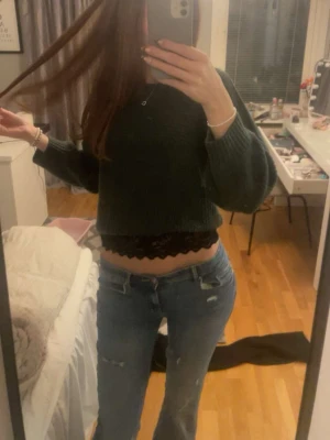 Mörkgrön croppad stickad tröja - Säljer en mörkgrön croppad stickad tröja med långa ärmar. Tröjan har en enkel, rak passform och ribbade muddar vid ärmslut och nederkant. Under tröjan syns en spetskantad topp som ger en snygg lager-på-lager-look, Nacken är klippt men fungerar fint använd fåtal gånger