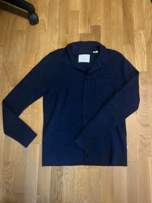 Mörkblå kofta från Jack & Jones - Säljer en mörkblå kofta från Jack & Jones med klassisk krage, knäppning framtill och en bröstficka. Koftan är långärmad och har en clean, stilren look som passar perfekt till lager-på-lager. Materialet är mjukt och stickat. Skriv vid frågor!