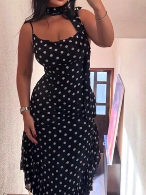 Svart prickig långklänning med volang - Säljer en svart långklänning med vita prickar och tunna axelband. Klänningen har en snygg volangkant nertill och medföljande scarf runt halsen. Figurnära passform och lätt v-ringad urringning ger en trendig look.