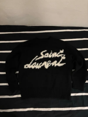 Svart Saint Laurent stickad tröja - Säljer en svart stickad tröja från Saint Laurent med ett stort vitt fuzzy logotyp-tryck på bröstet. Tröjan har rund hals, långa ärmar och breda ribbade muddar vid ärmslut och nederkant. Perfekt för dig som vill ha en statement piece i garderoben. Aldrig använd, hör av dig om du har några frågor.
