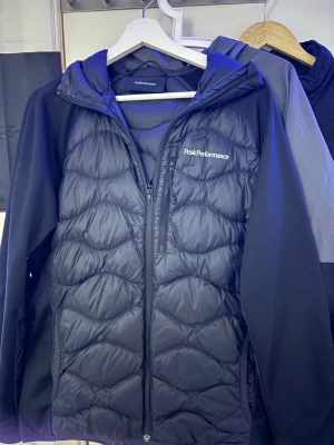 Svart pufferjacka Peak Performance L - Säljer en svart pufferjacka från Peak Performance i storlek L. Jackan har quiltad front, huva och dragkedja hela vägen. Ärmarna är släta och materialet är polyester. Perfekt för kyliga dagar och riktigt snygg streetstil.
