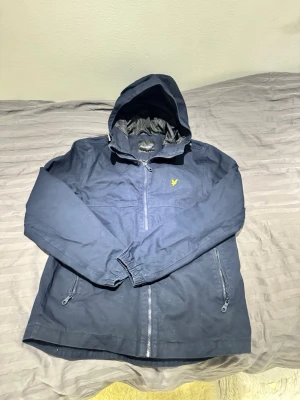Marinblå vindjacka från Lyle & Scott - Säljer en marinblå vindjacka från Lyle & Scott med huva och dragkedja framtill. Jackan har två fickor med dragkedja, elastiska muddar vid ärmsluten och den klassiska gula loggan på bröstet. Perfekt för blåsiga dagar och enkel att matcha med det mesta.